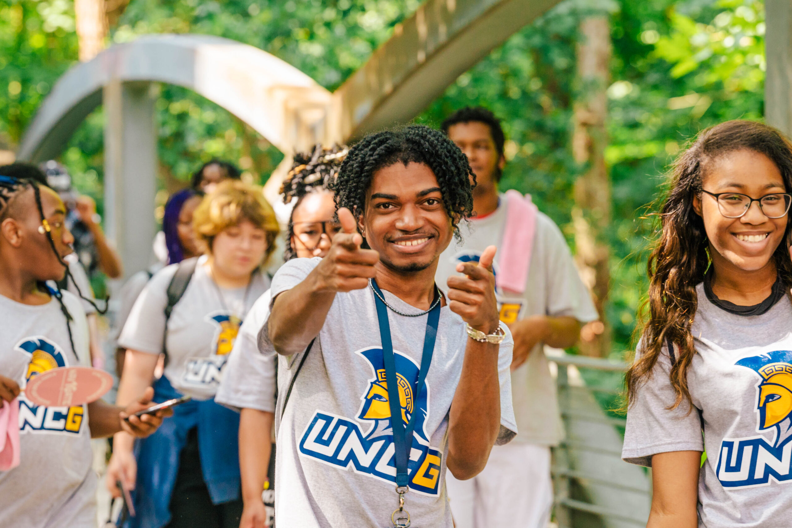 a group of students dressed in UNCG shirts, walking across a bridge together. The one in the center of the photo is looking at us and smiling, giving positive hand gestures.